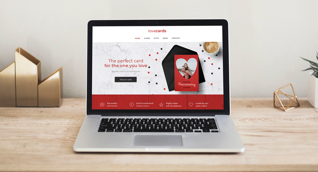 Laptop on a light wood desk with gold accessories. On the screen is a website for cards, gifts and ideas under the name Lovecards which fits the theme of St Valentine's Day. The theme of the site is red, white and black with a contemporary design.